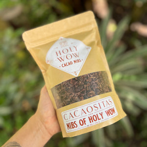 Hand holding a package of 'Nibs of Holy Wow' cacao nibs with a blurred natural background