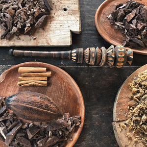 Wooden bowls with chocolate pieces, a wooden mallet, and a cacao pod on a dark surface.